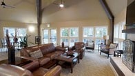 This inviting single-floor space is defined by its striking vaulted ceiling, which rises dramatically overhead and is supported by exposed structural beams that lend an architectural character to the room. A ceiling fan is centrally mounted, ensuring comfortable airflow throughout. The lounge area is anchored by a rich brown leather sectional sofa and coordinating armchairs, arranged thoughtfully around a solid wood coffee table — creating an atmosphere that is both welcoming and functional. Neutral-toned carpet adds warmth underfoot, complementing the earthy color palette of the walls and furnishings. A standout feature of this space is the generous arrangement of large white-framed windows and glass doors along the exterior wall, which bathe the interior in abundant natural light while offering clear views of lush greenery and an outdoor patio area beyond. Additional seating near the windows takes full advantage of these views, making the space feel seamlessly connected to the outdoors. The overall ambiance is cozy yet spacious, combining rustic and transitional design elements to create a comfortable communal gathering area suited for relaxation and social interaction.