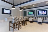 Located at 231 Market St in the heart of Alpharetta, GA, this single-floor amenity lounge is a refined communal space designed for both relaxation and social gathering. The interior is anchored by a striking central island clad in richly veined granite, topped with a matching stone countertop and outfitted with a sink — an ideal spot for casual entertaining, accompanied by stylish upholstered bar stools.

The ceiling features elegant tray detailing with bold black trim and recessed lighting, lending a polished, contemporary feel throughout. Pendant lights in a matte black finish hang above the central bar, adding a dramatic yet sophisticated touch. Light-toned large-format tile flooring runs seamlessly across the entire space, contributing to the bright and airy ambiance.

Multiple distinct seating zones are thoughtfully arranged throughout the room. A long upholstered banquette with framed flat-screen TVs creates a dedicated lounge and viewing area along one wall, while smaller tables and chairs are positioned for more intimate gatherings. A separate lounge nook features plush armchairs and geometric side tables, opening directly to an outdoor pool deck through full-height glass doors, flooding the space with natural light and offering seamless indoor-outdoor connectivity.
