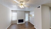 This single-floor residence offers a functional and well-appointed layout across several distinct areas. The living room serves as the heart of the home, anchored by a painted brick fireplace that adds warmth and character to the space. Rich wood-look flooring runs throughout the main living areas, complemented by a ceiling fan with a light fixture overhead and a textured popcorn ceiling. Natural light filters in through blinds-covered windows, and a glass-panel door provides access to the outdoors. A convenient pass-through opening connects the living area to the kitchen, which features white cabinetry and a practical countertop workspace. The bedroom is carpeted for comfort, with a large window that brings in additional natural light and neutral walls that create a calm, versatile atmosphere. The bathroom is clean and efficiently designed, featuring a full tub and shower combo with tiled surround, a toilet, white overhead cabinetry for storage, and a vanity topped with a speckled countertop and a framed mirror. An entry area with wood-look flooring and a paneled front door rounds out the layout. Throughout the space, a consistent neutral color palette and clean finishes provide a move-in-ready environment.
