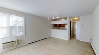 Located at 1202 Crescent Dr in Tarrytown, NY, this 2-bedroom, 1-bathroom unit offers a functional and comfortable single-floor layout. The open living area is finished with neutral wall-to-wall carpeting and light-toned walls, creating a bright and welcoming atmosphere. Natural light enters through a window fitted with horizontal blinds, complementing the track lighting overhead that illuminates the space effectively. A pass-through kitchen connects seamlessly to the living area, featuring warm wood-toned cabinetry, a white refrigerator, and a breakfast bar-style counter — a practical design that keeps the space feeling open and connected. The kitchen pass-through also includes an overhead light fixture, ensuring the cooking area is well-lit. A hallway visible from the main living space leads to the bedrooms and bathroom, providing a clear and efficient flow throughout the unit. Baseboard heating runs along the perimeter of the living room, offering a reliable climate control solution. The overall design is clean, straightforward, and move-in ready, making this Tarrytown residence a practical choice for those seeking comfortable living in a well-known Hudson Valley community.