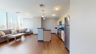 Located at 201 N Water St in downtown Wichita, Kansas, this one-bedroom, one-bathroom unit offers a well-organized layout across a single floor. The open-concept living, dining, and kitchen area forms the heart of the space, with warm hardwood-style flooring flowing throughout. The kitchen is equipped with stainless steel appliances, including a range, microwave, and refrigerator, complemented by light wood cabinetry and dark granite-style countertops. A central island with a dark countertop provides additional workspace and casual seating. Pendant lighting above the island adds a modern touch.

The dining area benefits from a structural column detail and large windows that allow ample natural light, with views of the downtown Wichita skyline visible from the living room's expansive window wall. A hallway connects the main living area to the private bedroom, which features plush dark carpeting and neutral gray walls, creating a quiet retreat. The bathroom includes a single-sink vanity with warm wood cabinetry, dark stone countertop, a large mirror, and a corner glass-enclosed shower. A dedicated closet completes the unit's practical layout.