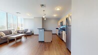 Located at 201 N Water St in downtown Wichita, Kansas, this one-bedroom, one-bathroom unit offers a well-organized layout across a single floor. The open-concept living, dining, and kitchen area forms the heart of the space, with warm hardwood-style flooring flowing throughout. The kitchen is equipped with stainless steel appliances, including a range, microwave, and refrigerator, complemented by light wood cabinetry and dark granite-style countertops. A central island with a dark countertop provides additional workspace and casual seating. Pendant lighting above the island adds a modern touch.

The dining area benefits from a structural column detail and large windows that allow ample natural light, with views of the downtown Wichita skyline visible from the living room's expansive window wall. A hallway connects the main living area to the private bedroom, which features plush dark carpeting and neutral gray walls, creating a quiet retreat. The bathroom includes a single-sink vanity with warm wood cabinetry, dark stone countertop, a large mirror, and a corner glass-enclosed shower. A dedicated closet completes the unit's practical layout.