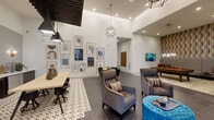 Located at Avalon Point in Missouri City, Texas, this single-story clubhouse is a thoughtfully designed communal space that blends contemporary style with comfortable functionality. Soaring ceilings throughout amplify the sense of openness, while floor-to-ceiling windows along one wall flood the interior with natural light and provide direct views of the surrounding outdoor deck and community.

The main lounge area anchors the space with a striking floor-to-ceiling stacked stone fireplace paired with a wall-mounted television, creating a warm focal point. Seating arrangements include plush armchairs, a vibrant orange sofa accented with teal pillows, and tufted ottomans, all arranged over large area rugs atop grey tile flooring. Geometric chandelier pendants and cage-style lanterns provide layered ambient lighting.

A dedicated wet bar and co-working zone features a tiled backsplash, counter seating, dark pendant lighting, and a distinctive slatted wood ceiling canopy overhead. Adjacent, a gallery wall of framed artwork adds a curated, editorial quality to the lounge corridor.

A games room with a pool table, retro-patterned accent wall, and its own chandelier provides entertainment, while decorative large-scale artwork and sculptural décor elements reinforce the space's polished, modern aesthetic throughout.