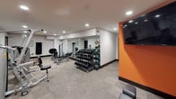 This dedicated fitness center is thoughtfully designed to support a comprehensive workout experience across a single open floor. The space is defined by a clean, modern aesthetic, with neutral carpet tiles underfoot, recessed lighting throughout, and bold orange accent walls that energize the environment.

The cardio zone is well-stocked with a lineup of Matrix brand equipment, including multiple treadmills, elliptical cross-trainers, and a recumbent stationary bike — each unit outfitted with an integrated digital display for interactive workouts. A central structural column anchors this zone and is flanked by a multi-station cable machine, ideal for a wide range of resistance exercises.

The strength and free weights area occupies a dedicated section of the room, featuring a full rack of dumbbells, a medicine ball collection, and a wall-length mirror that enhances the sense of space and aids in form monitoring. Additional selectorized strength machines line the perimeter, covering major muscle groups including chest press and leg extension stations.

A wall-mounted flat-screen television is positioned near the entrance, adding an entertainment element to the space. The overall layout is efficient and uncluttered, offering users both variety and functionality in a well-maintained, professional fitness environment.
