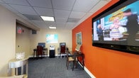 This single-floor space is designed to be both functional and visually engaging. The room makes an immediate impression with a striking orange accent wall that anchors the space and adds a bold, energetic character to the interior. Mounted prominently on this wall is a large flat-screen display, making it a natural focal point for presentations or media viewing.

The space is outfitted with multiple individual computer workstations, each paired with a chair, arranged to provide a comfortable and practical working environment. A dedicated desk near the display screen houses a tablet or touchscreen device, offering additional interactive functionality. A printer or multifunction device is also conveniently accessible within the room.

The flooring consists of patterned carpet tiles in a muted, multi-toned palette that complements the room's overall color scheme. Overhead, a drop ceiling with recessed panel lighting provides even, consistent illumination throughout the space.

Colorful framed artwork decorates the walls, adding a creative and welcoming atmosphere. The combination of practical workstations, multimedia capabilities, and thoughtful design details makes this a versatile space well-suited for business, education, or collaborative use.