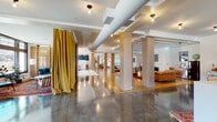 Located at 5193 Peachtree Rd in Chamblee, Georgia, this two-floor space masterfully blends industrial architecture with warm, eclectic interiors. The ground level features a mix of polished concrete and hardwood flooring throughout its expansive open areas, anchored by structural columns clad in light wood. A dramatic wall of floor-to-ceiling glass garage doors floods the main lounge with abundant natural light and offers views of the surrounding streetscape, while exposed ductwork and coffered white ceilings reinforce the space's industrial character. The lounge area is appointed with comfortable seating, open shelving with a large display TV, and pendant globe lighting that creates a welcoming ambiance. A communal dining area with a large farmhouse table, eclectic mixed chairs, and industrial swing-arm lighting sits beside black-framed windows overlooking an outdoor terrace. A hardwood-floored section features additional collaborative workspaces and enclosed offices with glass partitions. A distinctive slatted wood and glass partition wall adds architectural interest near a tiled entryway. The second floor, accessible via an open staircase, provides an overlooking mezzanine with additional seating and views down to the main floor. Throughout, vibrant Persian-style rugs add warmth and color against the cool industrial finishes, creating a cohesive and inviting environment.