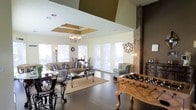 This single-floor residence showcases a thoughtfully designed open-concept layout that seamlessly blends living and entertainment spaces. The main living area is anchored by rich, dark hardwood flooring that extends throughout, creating a warm and cohesive aesthetic. A tray ceiling with elegant detailing adds architectural depth overhead, while a classic chandelier provides a refined focal point above the seating area.

Natural light floods the space through an array of large windows dressed with white horizontal blinds, offering a bright and airy atmosphere. The living area is generously proportioned, comfortably accommodating a full sofa and lounge seating arrangement centered around a glass-topped coffee table on a neutral area rug.

A striking exposed brick column serves as a bold architectural feature, complementing the deep brown accent wall and adding a touch of rustic character to the otherwise contemporary interior. An ornate wall medallion further enhances the decorative appeal of the space.

The entertainment area includes a foosball table, making it an ideal gathering space for recreation and socializing. A formal dining set with wrought iron chairs is also present, rounding out the versatility of this expansive, multi-functional open floor plan.