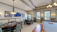 Located at 900 Grand Central Dr in Hamilton, NJ, this well-appointed game room offers a sophisticated yet relaxed atmosphere designed for recreation and socializing. The single-floor space is thoughtfully laid out to accommodate a variety of leisure activities without feeling cramped.

At the heart of the room sits a full-sized billiards table atop a warm hardwood floor platform, surrounded by neutral-toned carpeting that defines the surrounding seating areas. Multiple octagonal game tables with teal upholstered chairs are positioned throughout, ideal for card games or board games. Wall-mounted flat-screen TVs are strategically placed for easy viewing from multiple areas of the room.

Architecturally, the space features coffered and tray ceilings with crown molding and recessed lighting, lending a refined character to the room. Wainscoting with panel detailing lines the lower walls, complementing the neutral beige and sage green color palette. Pendant chandeliers and wall sconces provide warm, layered lighting throughout.

Natural light enters through multiple windows fitted with blinds, offering views of the surrounding outdoor greenery. High-backed bar-height chairs are positioned near the windows, creating additional casual seating. Framed artwork with automotive themes adds a distinctive decorative touch, completing the polished yet inviting ambiance of this community game room.