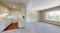 This single-floor unit is designed with a functional, straightforward layout that makes efficient use of its space. The living area is a notable highlight, featuring large windows that allow generous natural light to fill the room while offering views of the surrounding greenery and outdoor landscape. Neutral-toned carpeting extends throughout the living and bedroom areas, contributing to a cohesive, understated aesthetic. Baseboard heating is present throughout, ensuring consistent warmth across the unit.

The kitchen is partially separated from the living area by a dividing wall, creating a distinct cooking zone while maintaining an open connection to the main living space. It features hardwood-style flooring, white cabinetry, a ceiling-mounted range hood, and an electric coil range. A ceiling fan is positioned nearby, adding airflow to the adjoining area.

The bedroom is a comfortable, carpeted room with its own window offering outdoor views and natural light. A hallway connects the main living spaces to the bedroom and bathroom, maintaining a logical flow throughout.

The bathroom includes a vanity with white cabinetry, a wide mirror, mosaic tile flooring in muted tones, overhead cabinetry for storage, and baseboard heating. The overall color palette of the unit is light and neutral, creating a calm, move-in-ready environment.