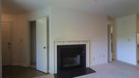 This single-floor residence offers a clean, understated aesthetic defined by neutral wall tones and plush carpeted flooring that lend the space a warm, welcoming atmosphere. The living area serves as the heart of the home, anchored by a classic fireplace framed with a crisp white mantel and contrasting dark surround and hearth. The fireplace adds both a functional focal point and a touch of timeless charm to the room. Vaulted or angled ceiling lines above the living area contribute to a sense of openness and architectural interest. The smooth, light-colored walls throughout provide a versatile backdrop well-suited to a variety of interior styles. Doorways visible from the main living area suggest a thoughtful layout with connected rooms offering comfortable flow between spaces. Subtle details such as electrical outlets and wall switches are neatly integrated into the walls, maintaining the clean lines of the interior. Overall, the space presents a straightforward, livable design with classic residential features that balance comfort and simplicity.