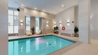 This 1,210 square foot indoor pool facility, located at 300 Place Charles-le Moyne in Longueuil, Quebec, is a testament to modern design and functionality. The space features a large, rectangular pool with crystal-clear turquoise water, complemented by a separate hot tub area. High ceilings and recessed lighting create an open, airy feel, while floor-to-ceiling windows flood the area with natural light and offer views of the surrounding cityscape. The pool deck is finished with light-colored tiles, enhancing the sense of cleanliness and space. Comfortable seating areas with potted plants add touches of warmth and greenery. Safety features include handrails, a life preserver, and clear signage. The facility also incorporates a changing area and appears to be wheelchair accessible, making it a welcoming space for all users. The overall design balances functionality with aesthetic appeal, creating a serene environment for swimming and relaxation.