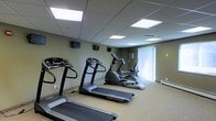 This single-floor fitness center offers a clean, functional environment designed for effective workouts. The space is outfitted with multiple treadmills, including Vision Fitness branded models, as well as elliptical trainers and a stationary bike, providing a well-rounded selection of cardio equipment for users of varying fitness levels. The room features a drop-panel ceiling with evenly spaced recessed lighting panels that illuminate the space with a bright, consistent glow. Wall-mounted monitors are positioned throughout the room, allowing users to stay entertained or informed during their workouts. The neutral-toned walls and light-colored floor contribute to a clean, open atmosphere. A window with vertical blinds along one wall allows for natural light to filter into the space, adding a sense of openness. The layout is thoughtfully arranged to provide ample room between machines, ensuring a comfortable and safe workout experience. Overall, this fitness center is a practical and well-maintained facility suited for regular cardiovascular training.