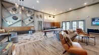 This thoughtfully designed single-floor amenity lounge offers a generous, open layout that seamlessly blends entertainment and social gathering spaces. The room is anchored by a striking feature wall finished in white brick, adorned with a geometric diamond-patterned panel surround and a large wall-mounted flat-screen television, complemented by a row of media storage cabinets beneath it. Recessed lighting and pendant fixtures illuminate the space, enhancing its modern, upscale ambiance.

The flooring transitions between distressed wood-look planks and a distinctive hexagonal tile pattern, visually defining different zones within the open layout. A centerpiece shuffleboard table sits prominently in the main gathering area, surrounded by rich cognac leather sofas and accent chairs that invite relaxed conversation. An angular built-in shelving unit with artistic décor adds a bold, sculptural element to one wall.

A communal high-top bar table with sleek black stools lines a padded banquette backed by warm brick, lit by gold sconce fixtures — ideal for social dining or casual meetings. A cozy windowed nook with built-in bench seating and a small round table provides a quieter retreat with views to the outdoors. French doors along one wall offer additional natural light and exterior access, completing this well-appointed, multifunctional lounge.