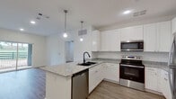 This well-appointed single-floor unit offers a clean, contemporary design highlighted by an open-concept layout that seamlessly connects the kitchen and living area. The kitchen serves as the focal point of the space, featuring crisp white shaker-style cabinetry, light granite countertops, and a central island with a black undermount sink. Pendant lighting above the island adds a stylish touch, while stainless steel appliances — including a refrigerator, electric range, over-the-range microwave, and dishwasher — complete the functional setup. Wood-look plank flooring extends throughout the entire unit, providing a cohesive and durable surface underfoot. The living area benefits from natural light and direct access to an outdoor patio through sliding glass doors, offering views of an open green landscape. The additional rooms include well-sized bedrooms, each with natural light from windows fitted with blinds. One bedroom features a ceiling fan for added comfort. Closet space is also present, providing practical storage. The overall palette of bright white walls, white trim, and neutral flooring creates a clean, modern aesthetic throughout the unit.