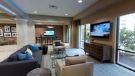 This well-appointed single-floor space offers a thoughtfully designed open-plan layout that seamlessly connects a comfortable lounge area with a dedicated dining space. The living area is furnished with a mix of plush sofas and accent chairs in neutral tones, centered around a glass coffee table and anchored by a large wall-mounted flat-screen television positioned above a sleek wooden media console. Large windows allow generous natural light to fill the room while offering views of lush greenery outside, creating a bright and welcoming atmosphere throughout.

A standout feature is the exposed brick accent wall that defines the dining area, lending a warm, urban character to the space. The dining section is outfitted with a dark-toned table and contemporary chairs, with a stylish pendant light overhead, making it ideal for both casual and more structured gatherings. Recessed lighting throughout the ceiling provides consistent, ambient illumination that complements the natural light. The flooring features a patterned area rug layered over neutral carpeting, adding texture and visual interest. The overall design palette—warm beiges, soft grays, and rich wood tones—creates a cohesive, modern aesthetic that is both functional and inviting.