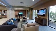 This well-appointed single-floor space offers a thoughtfully designed open-plan layout that seamlessly connects a comfortable lounge area with a dedicated dining space. The living area is furnished with a mix of plush sofas and accent chairs in neutral tones, centered around a glass coffee table and anchored by a large wall-mounted flat-screen television positioned above a sleek wooden media console. Large windows allow generous natural light to fill the room while offering views of lush greenery outside, creating a bright and welcoming atmosphere throughout.

A standout feature is the exposed brick accent wall that defines the dining area, lending a warm, urban character to the space. The dining section is outfitted with a dark-toned table and contemporary chairs, with a stylish pendant light overhead, making it ideal for both casual and more structured gatherings. Recessed lighting throughout the ceiling provides consistent, ambient illumination that complements the natural light. The flooring features a patterned area rug layered over neutral carpeting, adding texture and visual interest. The overall design palette—warm beiges, soft grays, and rich wood tones—creates a cohesive, modern aesthetic that is both functional and inviting.