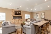 This single-story space is designed as an open, multi-functional gathering area that seamlessly integrates a fully equipped kitchen, a generous lounge, and dedicated dining zones. The layout flows naturally from one area to the next, creating a cohesive and welcoming environment throughout.

The kitchen is outfitted with dark espresso cabinetry, granite countertops with a stone tile backsplash, and stainless steel appliances including a wall oven, microwave, dishwasher, and full-size refrigerator. A large central island with bar-height seating provides additional prep space and casual dining options, while light tile flooring adds a clean, polished finish to this functional area.

The lounge area is a standout feature of the space, anchored by a dramatic floor-to-ceiling stacked stone accent wall that houses both a wall-mounted flat-screen television and a sleek linear electric fireplace. Vaulted ceilings with recessed lighting and soft neutral carpeting contribute to the warm, inviting ambiance. Multiple seating arrangements with gray upholstered sofas accommodate large groups comfortably.

The dining area includes a rustic wood table with mixed seating, positioned near windows that allow natural light and views of the surrounding community. Overall, the space presents a modern yet comfortable aesthetic suited for a variety of social occasions.