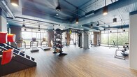 Located at 921 W First St in Winston-Salem, NC, this fitness center presents a thoughtfully designed, single-floor exercise facility that combines industrial aesthetics with comprehensive workout functionality. The space is characterized by its striking exposed ductwork painted in bold teal-blue tones, which runs throughout the high ceiling, complemented by warm brick accent walls and large floor-to-ceiling windows that flood the interior with natural light and offer views of the surrounding greenery and cityscape.

The main workout floor is generously sized and features durable wood-style flooring in the free weights and functional training zones, transitioning to carpet tile in the cardio area. A dedicated cardio section lines the perimeter with a full row of treadmills, ellipticals, and stair climbers. The functional training zone is anchored by a multi-use rig with TRX suspension straps and a heavy bag, supported by a well-stocked rack of medicine balls, kettlebells, and battle ropes. The strength area includes cable machines, weight benches, and barbells with ample open floor space for stretching and movement. A separate, glass-partitioned studio room with mirrored walls offers space for group classes or personal training. A built-in cubby station provides convenient storage for personal belongings.
