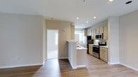 This single-floor residence offers a thoughtfully designed layout centered around an open-concept kitchen and living area. The kitchen is well-appointed with stainless steel appliances, including a range, microwave, and refrigerator, complemented by light wood cabinetry that provides ample storage. A white peninsula with an integrated sink serves as both a functional workspace and a casual dining or gathering spot, seamlessly connecting the kitchen to the adjoining living area.

Warm hardwood-style flooring runs throughout the space, lending a cohesive and inviting aesthetic. Recessed lighting in the kitchen ensures bright, even illumination, while multiple windows allow natural light to filter in, creating an airy atmosphere. The neutral beige and white wall tones throughout enhance the sense of openness.

A doorway off the main living area leads to additional spaces within the unit, offering a practical flow between rooms. The clean, contemporary finishes and efficient use of space make this residence a comfortable and functional home suited for everyday living.
