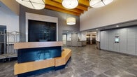 Located at 101 Summer St in Stamford, Connecticut, this ground-floor lobby presents a polished and professionally designed entrance experience. The space is anchored by a distinctive angular reception desk crafted from warm wood tones with a dark base, positioned beneath dramatic exposed wood beam ceiling accents and oversized drum pendant lighting that lend the area an architectural character. A large decorative dark-tile water wall feature serves as a striking focal point adjacent to the reception area, adding a sense of sophistication to the environment.

The flooring throughout is finished in large-format stone-look tile in a neutral gray tone, providing a durable yet refined foundation. A dedicated lounge area offers white upholstered seating arranged around a glass-top coffee table, accented by a vibrant green area rug and floor-to-ceiling drapes framing a large window that draws in natural light and views of the streetscape.

Additional functional amenities include a bank of modern package lockers for convenient parcel management, a glazed concierge or management office visible from the main corridor, and a set of stainless steel elevators serving the building. The entry vestibule features glass double doors with a building identification mat, completing the professional and welcoming first impression this lobby is designed to deliver.