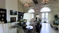 This single-floor space exudes refined elegance through its thoughtful architectural design and carefully curated interiors. The main living area commands attention with dramatic vaulted ceilings accented by dark exposed wooden beams, creating a striking visual contrast against the crisp neutral walls. Arched windows flood the space with abundant natural light, while French doors open to an outdoor area, seamlessly blending interior and exterior living. Two stately chandeliers with a candlestick-style design hang overhead, casting a warm, inviting glow throughout. A classic fireplace anchors one wall, flanked by dark built-in shelving that offers both display space and functional storage, alongside a mounted flat-screen television. The seating arrangement is generously appointed with a plush sofa, striped armchairs, and a mix of dark-finished side and console tables that lend a grounded, contemporary feel. Statement table lamps with bold white bases add a modern touch, while lush greenery and floral arrangements throughout the space bring life and warmth. The overall ambiance balances traditional architectural character with modern furnishings, resulting in a space that feels both grand and comfortably livable.