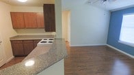 This single-floor space showcases an open-concept layout that seamlessly connects the kitchen and living area, creating a cohesive and functional environment. The kitchen is equipped with a white electric cooktop set into granite-style countertops that extend along a peninsula, offering additional workspace and a natural divider between the kitchen and the adjacent living area. Rich, warm-toned wood cabinetry lines the upper walls of the kitchen, providing ample storage while adding a traditional aesthetic to the space. The flooring throughout is a medium-toned hardwood, lending warmth and continuity as it flows from the kitchen into the open living area. The living space is bright and open, with neutral sage-green walls that create a calm, inviting atmosphere. A ceiling fan is visible overhead, contributing to the room's practicality and comfort. Clean white baseboards frame the flooring and provide a crisp contrast to the walls. A doorway visible in the background hints at additional rooms beyond the main living space. Overall, the layout and finishes combine to create a comfortable, move-in-ready environment with a classic, understated design sensibility.