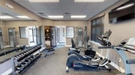 This fitness center is a comprehensively outfitted facility designed to support a wide range of workout routines. The space is organized across a single floor and divided into functional zones, each tailored to a specific type of exercise.

The main workout area features a selection of Diamondback cardio equipment, including elliptical machines and an upright stationary bike, positioned along the walls beneath a wall-mounted television. A full cable resistance machine provides options for strength training, while a dumbbell rack stocked with a range of free weights and an accompanying flat bench supports traditional weightlifting. Large mirrors line one wall, enhancing the sense of openness throughout the space.

A secondary area serves as a dedicated stretching and functional fitness zone, equipped with a colorful set of stability balls in multiple sizes, a water cooler station, and sanitation supplies for convenience. Stylish dark-framed glass doors connect the zones, adding a refined architectural detail.

Throughout, the flooring consists of durable rubber-speckled carpet suited for gym use, and the drop-panel ceiling is fitted with recessed fluorescent lighting. Multiple windows dressed with horizontal blinds allow natural light while maintaining privacy, contributing to a clean, neutral, and functional ambiance.