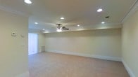 This single-floor space presents a clean, well-maintained interior designed with both comfort and functionality in mind. The main room is generously proportioned, offering an open and versatile layout that can accommodate a variety of uses. The ceiling is finished with elegant crown molding and equipped with multiple recessed lights that provide bright, even illumination throughout the space. A central ceiling fan adds both aesthetic appeal and practical air circulation. The walls are painted in a neutral, light tone that enhances the sense of openness and pairs well with the plush wall-to-wall carpeting underfoot, which adds warmth and softness to the room. Vertical blinds on the window allow for adjustable natural light control, letting in soft, diffused daylight while maintaining privacy. White baseboard trim runs along the perimeter of the room, adding a crisp, finished look to the overall interior. The space is move-in ready and presents a blank canvas ideal for customization, whether as a living area, recreational room, or multipurpose space.