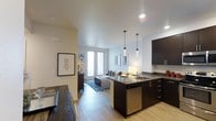 This single-floor space showcases a thoughtfully designed open-concept layout that integrates the kitchen, dining, and living areas into one cohesive and functional environment. The kitchen is a standout feature, outfitted with rich dark cabinetry, dark countertops, and stainless steel appliances, including a dishwasher. A peninsula-style counter with an undermount sink serves as both a prep area and a natural divider between the kitchen and the rest of the space. Three sleek pendant lights hang above, adding a modern, refined touch to the area. Light hardwood flooring runs throughout, lending warmth and continuity to the interior. The dining area sits adjacent to the kitchen, furnished with a dark wood table and coordinating chairs. The living area beyond is bright and welcoming, benefiting from generous natural light streaming through large glass doors or windows at the far wall. The living space is styled with a comfortable sofa, decorative throw pillows, and a patterned area rug that anchors the seating arrangement. Clean white walls, recessed lighting, and tasteful wall art contribute to the modern, contemporary ambiance that defines this well-appointed space.