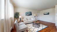 This single-floor residence offers a thoughtfully organized layout that balances comfort and functionality across its main living areas. The living room serves as the heart of the home, showcasing rich hardwood flooring, neutral walls, and generous natural light filtering through sheer curtains. The space comfortably accommodates a full seating arrangement and flows naturally into a dedicated dining area, which features the same warm hardwood floors, a pendant light fixture centered above a round dining table, and a window with a wall-mounted air conditioning unit.

The kitchen is efficiently arranged with warm wood cabinetry, stone-look countertops, stainless steel appliances, and durable tile flooring, providing ample workspace and storage. Two bedrooms offer carpeted and hardwood flooring options respectively, each with windows and sufficient space for full bedroom furniture. The primary bedroom includes direct access to a bathroom featuring warm wood vanity cabinetry, a white countertop, and a tiled tub-shower combination. A second full bathroom mirrors this design with matching tile flooring and a vanity, maintaining a cohesive aesthetic throughout the residence.