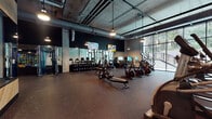 This fitness center combines industrial aesthetics with a fully equipped workout environment. The space features soaring exposed concrete ceilings with visible ductwork and conduit, pendant lighting suspended from above, and floor-to-ceiling windows along one wall that flood the interior with natural light and provide views of surrounding greenery. The rubber flooring throughout offers durability and safety across all workout zones.

The strength training area is anchored by a full rack of dumbbells displayed along a mirrored wall, accompanied by adjustable benches and cable machines. Large mirrors extend across multiple walls, visually expanding the space and allowing users to monitor their form. Warm wood-paneled accent walls and built-in storage benches provide contrast against the dark, moody color palette of charcoal and deep teal.

The cardio zone is well-stocked with Matrix-brand treadmills, ellipticals, stair climbers, and stationary cycling bikes, several of which are equipped with individual display screens. Mounted flat-screen televisions are positioned throughout for entertainment. The open layout ensures generous spacing between equipment, creating a comfortable and functional training environment.