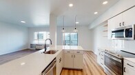 This thoughtfully designed single-floor residence showcases a clean, modern aesthetic carried consistently throughout every room. The open-concept kitchen serves as the heart of the home, featuring crisp white cabinetry with matte black hardware, a quartz waterfall countertop, and a central island with an undermount stainless steel sink. Stainless steel appliances — including a range, over-the-range microwave, and refrigerator — complement the subway tile backsplash, while warm-toned floating wood shelves add a touch of natural contrast. Pendant lighting and recessed ceiling lights illuminate the space, which flows seamlessly into the adjoining living area. Light-toned hardwood-style flooring runs throughout, lending warmth and visual continuity to the entire residence. The bedrooms are bright and airy, each benefiting from generous natural light through large windows that frame views of the surrounding neighborhood. The primary bathroom features a wide quartz vanity, ample under-sink storage, a large frameless mirror, and matte black fixtures, while the secondary bathroom offers a full tub-shower combination with coordinating black hardware. The overall ambiance is fresh, clean, and move-in ready, with a minimalist design palette that provides a versatile canvas for any style.