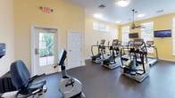 This fitness center offers a comprehensive workout environment spread across a single floor, thoughtfully designed to accommodate a variety of exercise needs. The space features warm yellow and neutral-toned walls, durable rubber flooring, and a combination of flush-mount and pendant lighting, creating a clean and energizing atmosphere.

The main cardio area is well-stocked with TRUE-brand treadmills, elliptical trainers, and stationary bikes, arranged to maximize floor space and allow comfortable circulation between equipment. Clerestory windows along the upper walls, along with larger windows and a glass-paneled exterior door, invite generous natural light and outdoor views throughout this section of the space.

A ceiling fan enhances air circulation, contributing to a comfortable workout environment. A wall-mounted television provides entertainment during exercise sessions.

A secondary area houses a dumbbell rack with a full range of free weights, along with adjustable workout benches and a recumbent bike, catering to strength and low-impact training. A dedicated nook with a children's table and chairs makes the facility family-friendly, allowing parents to exercise while keeping an eye on young children nearby.