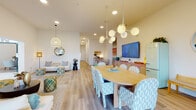 This single-floor space offers a thoughtfully designed open layout that seamlessly connects multiple functional areas, creating a bright and inviting atmosphere throughout. Light wood flooring extends across the entire space, unifying the various zones while contributing to the warm, airy feel.

The entrance area makes a strong architectural statement with soaring ceilings and distinctive arched doorways framed by large glass panels, flooding the foyer with natural light. A round console table anchors this entry vestibule, setting a welcoming tone.

The lounge area features a cream scallop-edged sofa, houndstooth ottomans, a round coffee table, and a tiered shelving unit displaying curated décor. Arched windows allow additional natural light, while a dedicated workstation with a desk and seating provides a functional workspace nearby.

The dining area is a standout, centered around a large oval wood table surrounded by upholstered chairs in a light blue geometric pattern. A sculptural multi-globe pendant light hangs overhead. A pastel pink cane sideboard, a retro mint-green refrigerator, and bold wall art — including a large rainbow graphic piece — add playful, eclectic character.

Throughout, the space balances vibrant color accents with clean white walls, resulting in a modern, cheerful, and highly functional environment.