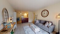 This efficiently designed single-floor unit offers a thoughtful layout encompassing a living room, kitchen, bedroom, closet, and hallway. The open-concept living area is warm and inviting, finished with soft wall-to-wall carpeting and neutral cream-toned walls that create a bright, cohesive atmosphere. A ceiling fan overhead provides comfortable airflow, while a large sliding glass door floods the space with natural light and provides direct access to a private outdoor patio with views of the surrounding community. The kitchen is positioned adjacent to the living area and features warm wood cabinetry, light countertops, a full-size refrigerator, and a raised breakfast bar with seating — ideal for casual dining. Tile flooring in the kitchen adds a practical contrast to the carpeted living spaces. A dedicated dining area sits comfortably near the kitchen, enhanced by a ceiling fan with integrated lighting and recessed fixtures above. The bedroom is a serene retreat, carpeted and well-lit by a window dressed with teal curtains that invite natural light while maintaining privacy. A ceiling fan completes the room. The closet provides convenient storage, and the hallway connects all areas in a smooth, functional flow throughout the unit.