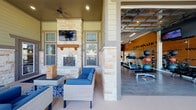 Located at 11211 Westwood Loop in San Antonio, Texas, this fitness center and outdoor lounge facility offers a comprehensive wellness environment designed for community use. The interior gym space features wood-look flooring, vaulted ceilings with pendant and ceiling fans, and bold orange accent walls adorned with motivational typographic murals. The facility is equipped with a full array of cardio machines, including treadmills, stair climbers, and stationary bikes, each outfitted with individual media screens. A dedicated free weights area includes a tiered dumbbell rack, kettlebell sets, stability balls, and a workout bench, complemented by floor-to-ceiling mirrors that amplify the sense of space. Natural light floods the gym through large glass panel garage-style doors that open directly to the outdoor patio, seamlessly blending indoor and outdoor environments. The covered outdoor lounge area is anchored by a striking floor-to-ceiling stacked stone fireplace with a wood mantel and a mounted flat-screen television above. Comfortable wicker seating with blue cushions invites relaxation, while recessed lighting and a ceiling fan ensure year-round comfort. The covered patio's stone and lap siding exterior detailing adds architectural character, with views of the surrounding community visible beyond the iron railing perimeter.