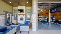 Located at 11211 Westwood Loop in San Antonio, Texas, this fitness center and outdoor lounge facility offers a comprehensive wellness environment designed for community use. The interior gym space features wood-look flooring, vaulted ceilings with pendant and ceiling fans, and bold orange accent walls adorned with motivational typographic murals. The facility is equipped with a full array of cardio machines, including treadmills, stair climbers, and stationary bikes, each outfitted with individual media screens. A dedicated free weights area includes a tiered dumbbell rack, kettlebell sets, stability balls, and a workout bench, complemented by floor-to-ceiling mirrors that amplify the sense of space. Natural light floods the gym through large glass panel garage-style doors that open directly to the outdoor patio, seamlessly blending indoor and outdoor environments. The covered outdoor lounge area is anchored by a striking floor-to-ceiling stacked stone fireplace with a wood mantel and a mounted flat-screen television above. Comfortable wicker seating with blue cushions invites relaxation, while recessed lighting and a ceiling fan ensure year-round comfort. The covered patio's stone and lap siding exterior detailing adds architectural character, with views of the surrounding community visible beyond the iron railing perimeter.