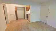 This single-floor unit offers a straightforward and functional layout designed for comfortable everyday living. The main living area is finished with wall-to-wall carpeting in a neutral beige tone, creating a warm and cohesive atmosphere throughout the space. A large built-in closet with classic bifold doors provides generous storage directly off the living area, contributing to an organized and clutter-free environment. A floor vent is integrated into the wall, ensuring efficient climate control throughout the unit. Adjacent to the living space, the kitchen is partially open to the main area, featuring warm wood-toned upper and lower cabinetry that adds a traditional character to the space. The kitchen is equipped with a sink and a dishwasher, offering practical amenities for daily use. The walls throughout are finished in a clean, light neutral tone that enhances the sense of brightness and openness. A secondary door is visible along the far wall, suggesting additional access or storage options. Overall, the unit presents a clean, move-in-ready interior with a simple, utilitarian design suited for a range of occupants.
