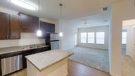 This single-floor residence offers a thoughtfully designed open-concept layout that seamlessly connects the kitchen and living area, creating a spacious and welcoming atmosphere. The kitchen is appointed with rich dark cabinetry, granite-look countertops, and stainless steel appliances, including a full-size refrigerator. A peninsula with a breakfast bar provides additional prep space and casual seating, while two pendant lights hang above, adding a warm, modern touch. A flush-mounted light fixture further brightens the space. The kitchen flows naturally into the living area, which transitions from hardwood-style flooring to plush carpeting, defining the two functional zones. The living area is bathed in natural light from a large multi-panel window, and a ceiling fan overhead ensures year-round comfort. Neutral wall tones and clean lines throughout contribute to a bright, airy ambiance. The overall design balances practicality with contemporary style, making the space both functional and inviting for everyday living.