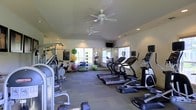 This fitness center offers a comprehensive workout environment designed to accommodate a range of exercise routines. The space is generously proportioned, with a layout that allows for comfortable movement between equipment zones. Two ceiling fans help maintain air circulation throughout the room, complementing the recessed lighting overhead.

The cardio area is prominently featured along one side of the room, housing multiple treadmills and elliptical machines, each equipped with individual monitors for entertainment or workout tracking. A wall-mounted television is also positioned for shared viewing. Large windows along the back wall allow natural light to fill the space, creating a bright and motivating atmosphere while offering views of the surrounding greenery.

On the opposite end, a dedicated functional training area features stability balls in various sizes, resistance equipment, and additional strength training machines. A mirrored wall in this section enhances the sense of space and allows users to monitor their form during workouts. The flooring throughout consists of durable, gray carpet tiles suited for high-traffic fitness use. Overall, the space presents a clean, organized, and well-maintained environment suitable for a full workout regimen.