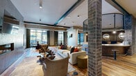 This generously sized single-floor amenity space combines industrial character with refined design across an open, flowing layout. Reclaimed hardwood flooring runs throughout, anchoring the warm yet contemporary aesthetic. Exposed steel beams and structural brick columns add architectural depth, while the metallic textured accent wall serves as a dramatic focal point behind the lounge's built-in linear fireplace and wall-mounted TV.

At the heart of the space sits a striking curved bar with a patinated metal façade and a light stone countertop, encircled by a decorative industrial mesh canopy overhead — a true centerpiece for social gatherings. Adjacent lounge seating areas are arranged around area rugs and eclectic furnishings, creating inviting pockets for relaxation or conversation.

A dedicated dining area features a dark round table set before expansive floor-to-ceiling windows, framing views of an outdoor terrace and courtyard. Elegant drape panels soften the natural light that floods the space throughout the day. Large-scale abstract artwork adorns the walls, lending a curated gallery quality to the environment.

The entry corridor features clean tile flooring, circular pendant lighting, and a reception-style package station. A fitness area rounds out the space's offerings, making this a fully equipped and visually cohesive community hub.