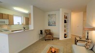This single-floor residence offers a thoughtfully designed layout that balances openness with functional detail. The living area serves as the heart of the home, featuring soft wall-to-wall carpeting that adds warmth underfoot and neutral, light-toned walls that create a welcoming, airy atmosphere. A standout architectural element is the built-in shelving unit integrated into a structural column, providing both a decorative display space and a subtle room divider that adds character to the layout. A nautical-themed art piece adorns the main wall, contributing to the space's calm, coastal aesthetic. The living area flows naturally toward the kitchen, which is accessible via a pass-through window — a practical design choice that maintains visual connectivity between the two spaces while preserving distinct functional zones. The kitchen features light wood cabinetry and a bright overhead fixture, keeping the cooking area well-lit. Throughout the space, recessed and flush-mount lighting ensures consistent illumination. The overall ambiance is clean and understated, with a design sensibility that emphasizes simplicity, natural light, and versatile living — making it a comfortable and practical single-floor home.