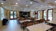 This single-floor community room serves as a versatile dining and social gathering space. The interior is defined by a coffered ceiling with recessed lighting, which lends a structured, polished aesthetic to the room. Light-toned tile flooring runs throughout, contributing to a clean and open atmosphere.

Multiple rectangular tables are arranged throughout the space, each surrounded by upholstered chairs featuring a warm floral pattern, providing comfortable seating for a considerable number of occupants. The furniture arrangement encourages both communal dining and casual gatherings.

Along the back wall, built-in dark wood shelving units flank a wall-mounted flat-screen television, suggesting the space doubles as a media or lounge area. The shelving appears to hold books and decorative items, adding a functional and homey element to the room.

Large glass doors line the perimeter walls, flooding the interior with natural light and offering views of a landscaped outdoor area, visually connecting the inside to the exterior environment. The overall ambiance is warm, welcoming, and well-suited for community activities and shared dining experiences.