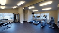 This single-floor fitness center is thoughtfully designed to provide a functional and comfortable workout environment. The space is outfitted with durable dark rubber flooring, offering both safety and practicality for a variety of exercise activities. Bright flush-mounted rectangular ceiling lights ensure the room is evenly and generously illuminated throughout.

Along one wall, a row of treadmills and an elliptical machine are positioned in front of large windows that invite natural light and offer views of the surrounding greenery, creating an energizing atmosphere for cardio workouts. A wall-mounted flat-screen television is positioned above the windows, adding entertainment options for users during their sessions.

On the opposing side of the room, a multi-station cable and weight machine provides users with a wide range of strength training options. Mirrors are integrated into this area, enhancing the sense of space while allowing users to monitor their form during exercises.

The room features neutral-toned walls with clean white trim and a paneled door, contributing to a tidy and organized aesthetic. A ventilation grille is visible near the ceiling, ensuring proper air circulation throughout the space. Overall, the fitness center strikes a balance between practicality and comfort, making it a well-rounded facility for daily exercise routines.