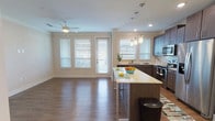 This well-appointed single-story unit offers a thoughtfully designed layout that maximizes functionality and comfort. The open-concept living and kitchen area serves as the heart of the home, where rich hardwood-style flooring flows continuously underfoot and crown molding adds a refined architectural detail. A ceiling fan overhead enhances the airy feel, while multiple windows fitted with horizontal blinds invite abundant natural light and offer outdoor views.

The kitchen is a standout feature, showcasing dark espresso cabinetry paired with a crisp light countertop, a center island with seating, stainless steel appliances including a refrigerator, range, microwave, and dishwasher, and stylish pendant lighting above the island. Recessed lighting complements the pendants to ensure a well-lit workspace.

The bedroom is generously proportioned, featuring a ceiling fan, natural light from dual windows, and direct access to a walk-in closet with built-in shelving. Crown molding continues the refined detailing found throughout the unit.

The bathroom presents a clean, neutral palette with large-format tile flooring, a tiled tub-shower surround, and a dark-toned vanity with a stone-look countertop.

A dedicated laundry room with a full-size washer and dryer and overhead wire shelving rounds out this practical and polished living space.