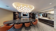 This elegantly designed single-floor space functions as a premium meeting and lounge facility. The centerpiece of the conference area is a large, polished dark granite table surrounded by cognac leather chairs with slender metal legs, accommodating a sizeable group for meetings or events. Overhead, a dramatic sculptural chandelier composed of clustered circular diffusers serves as an artistic focal point, casting warm, ambient light throughout the room.

The walls are framed by dark wood built-in shelving units, backlit to highlight curated decorative objects including ceramics, vases, and sculptural accents. A wall-mounted flat-screen television is positioned on a textured accent wall, making the space equally suited for presentations. Patterned broadloom carpeting in neutral tones runs throughout, adding texture underfoot.

Glass sliding partitions with brass-toned hardware allow sections of the space to be separated or opened, providing flexible configurations. Adjacent areas include a lounge zone with additional seating, a round table with short stools, and a reception-style counter, all connected visually through the transparent partitions. Natural light filters in through frosted windows, complementing the warm artificial lighting throughout.