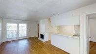 This single-floor residence offers an open-concept layout that seamlessly connects the living area and kitchen. The living space is anchored by a classic white-painted fireplace, serving as a central focal point, and is finished with warm hardwood flooring that extends throughout. A large window near the front entry fills the room with natural light, offering a view of an outdoor balcony railing, adding a sense of openness to the space. The textured popcorn ceiling adds a traditional character overhead, while HVAC venting ensures year-round climate comfort. The kitchen features a wraparound breakfast bar with a white countertop, providing both functional workspace and casual seating potential. White cabinetry lines the upper portion of the kitchen area, offering ample storage. A hallway visible from the main living area leads to additional rooms, suggesting a practical and well-organized floor plan. The overall aesthetic is clean and neutral, with an all-white palette across the walls, cabinetry, and fireplace surround, complemented by the warmth of the wood flooring — creating a bright, welcoming environment.