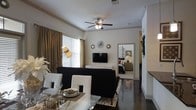 This single-floor residence showcases a thoughtfully designed open-concept layout that connects the dining and living areas in a seamless, flowing arrangement. The interior is anchored by rich, dark hardwood floors that extend throughout, providing a striking contrast against the crisp white walls and ceiling. A ceiling fan with integrated lighting adds both function and a classic touch overhead.

The dining area features a glass-top table paired with sleek white chairs, creating an elegant yet contemporary setting. Adjacent to it, the living area is furnished with a bold black sofa and complementary accent pieces, centered around a wall-mounted flat-screen television atop a white console. Decorative sunburst wall art adds a stylish focal point to the space.

Natural light filters in through large windows dressed with warm golden drapes and horizontal blinds, enhancing the bright, welcoming ambiance. A glimpse through a framed opening reveals a well-appointed bedroom beyond, suggesting a connected and cohesive floor plan. The overall aesthetic blends modern and transitional design elements, resulting in a polished, comfortable living environment with a warm yet sophisticated character throughout.