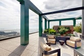 Located in San Francisco's Mission Bay neighborhood, this rooftop terrace at 1070 Bridgeview Way offers a striking outdoor experience defined by architectural boldness and panoramic urban scenery. The terrace is anchored by a series of distinctively colored green steel pergola columns and overhead beams that frame the open sky, creating a visually dynamic structure throughout the space. Underfoot, speckled terrazzo-style tiles extend across the generous terrace floor, lending a refined, durable finish suited to the outdoor environment. Glass panel railings line the perimeter, ensuring unobstructed views of San Francisco Bay, the Bay Bridge, the city skyline, and the waterfront below. Curated seating areas are thoughtfully arranged beneath the pergola, featuring contemporary lounge chairs, ottomans, and side tables that invite relaxed gatherings. Raised planter beds filled with lush, drought-tolerant greenery and ornamental plants add natural texture and color. Additional casual dining furniture, including bistro-style tables and chairs in a warm red tone, provide flexible spaces for outdoor dining or informal meetings. The corrugated metal cladding of the rooftop mechanical structure complements the industrial-modern aesthetic of the building. This rooftop terrace offers a compelling combination of design, comfort, and some of San Francisco's most captivating views.