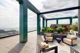 Located in San Francisco's Mission Bay neighborhood, this rooftop terrace at 1070 Bridgeview Way offers a striking outdoor experience defined by architectural boldness and panoramic urban scenery. The terrace is anchored by a series of distinctively colored green steel pergola columns and overhead beams that frame the open sky, creating a visually dynamic structure throughout the space. Underfoot, speckled terrazzo-style tiles extend across the generous terrace floor, lending a refined, durable finish suited to the outdoor environment. Glass panel railings line the perimeter, ensuring unobstructed views of San Francisco Bay, the Bay Bridge, the city skyline, and the waterfront below. Curated seating areas are thoughtfully arranged beneath the pergola, featuring contemporary lounge chairs, ottomans, and side tables that invite relaxed gatherings. Raised planter beds filled with lush, drought-tolerant greenery and ornamental plants add natural texture and color. Additional casual dining furniture, including bistro-style tables and chairs in a warm red tone, provide flexible spaces for outdoor dining or informal meetings. The corrugated metal cladding of the rooftop mechanical structure complements the industrial-modern aesthetic of the building. This rooftop terrace offers a compelling combination of design, comfort, and some of San Francisco's most captivating views.